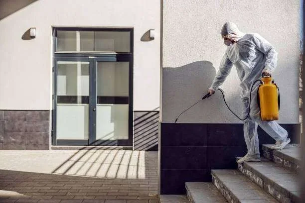 Profissional com EPI e pulverizador amarelo desinfetando escadas e parede de edifício